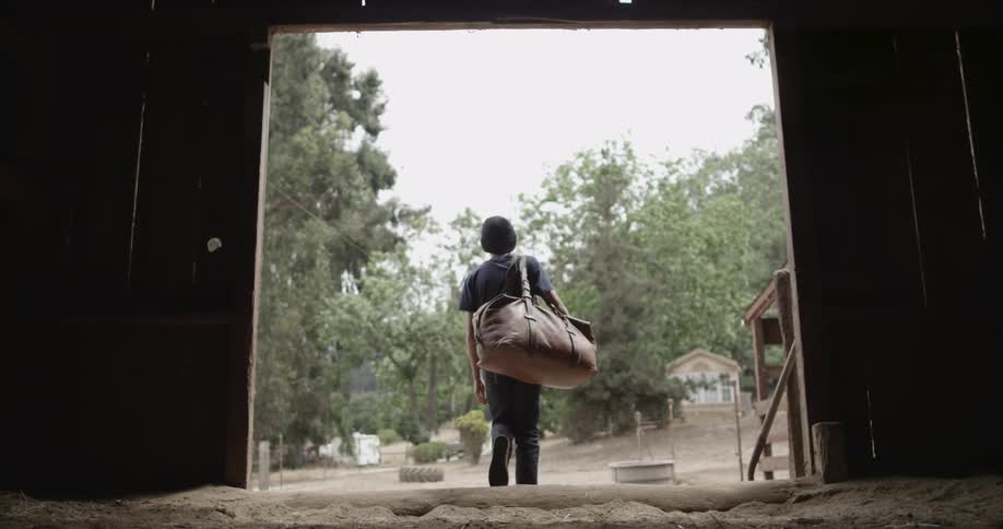 Boy holding a bag walks out of a barn on a cloudy day