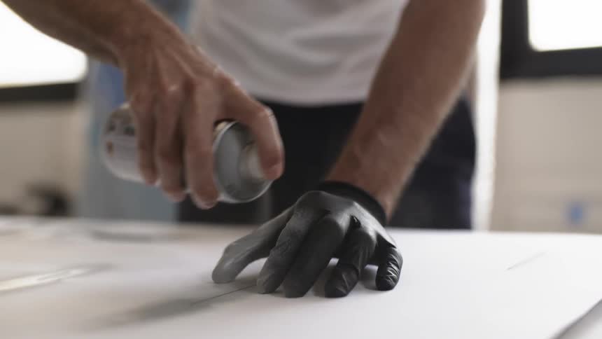 A man spray paints an object on a tool bench and his gloved hand