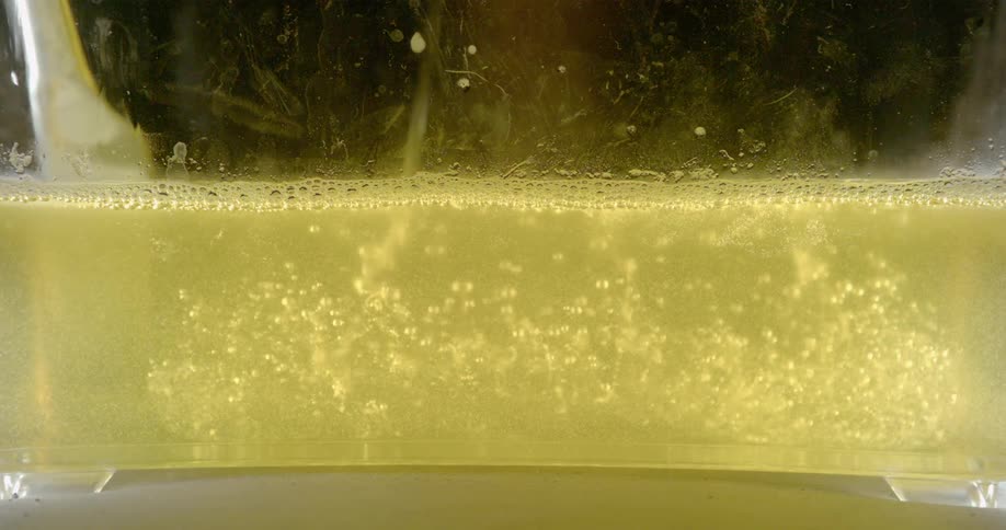Liquid is poured into a clear container that already has yellow liquid in it and bubbles float up