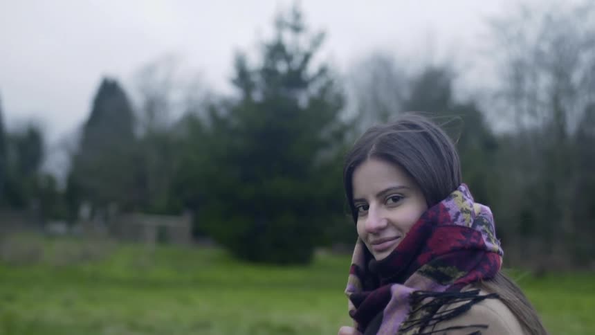 Woman walking through a field and moving a scarf around her neck while looking at the camera