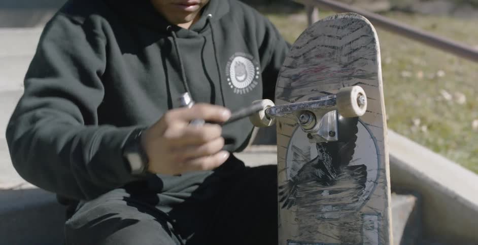 A male in a black sweatshirt uses a tool to tighten the center of the truck at one of his skateboard