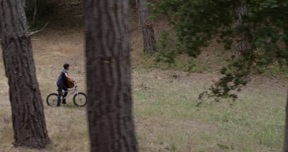A boy carrying a large bag rides a bike through a field