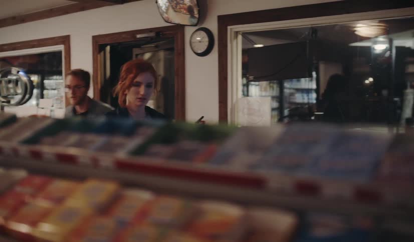 A red headed woman and bespectacled male enter a convenience store