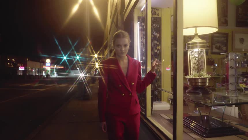 A woman in a pink suit walks in front of a store window at night