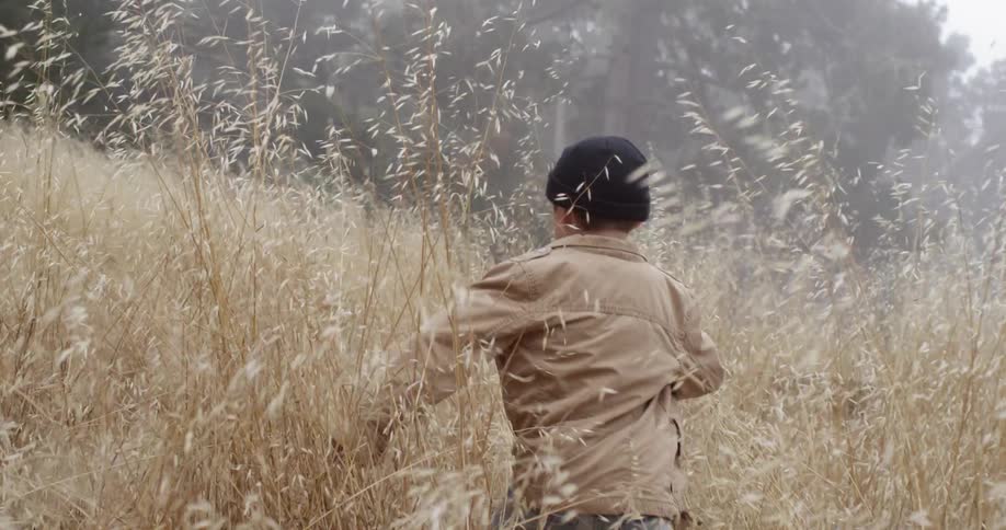 Boy running through a tall grass field towards trees on a cloudy day