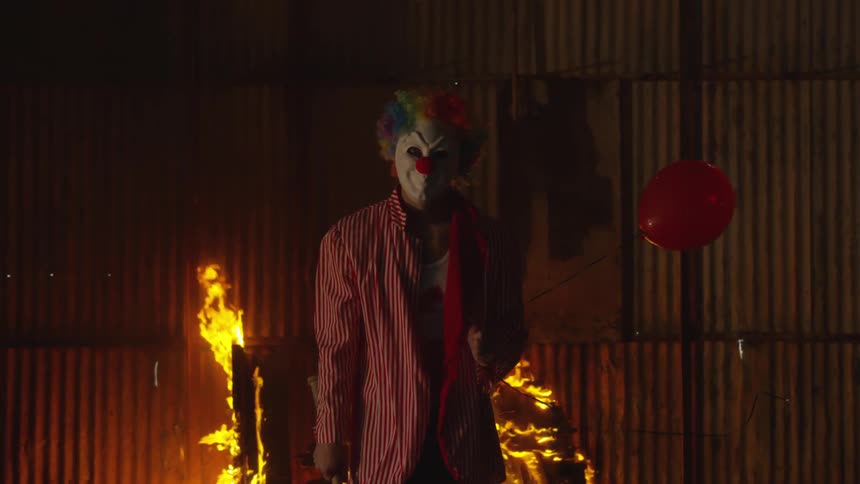 A person dressed as a clown is by a barn on fire