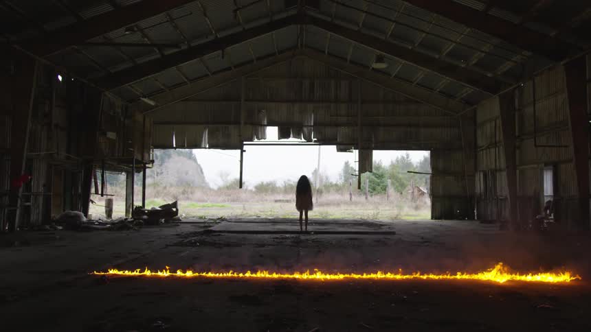 A young girl stands in an abandoned building while a fire burns on the ground