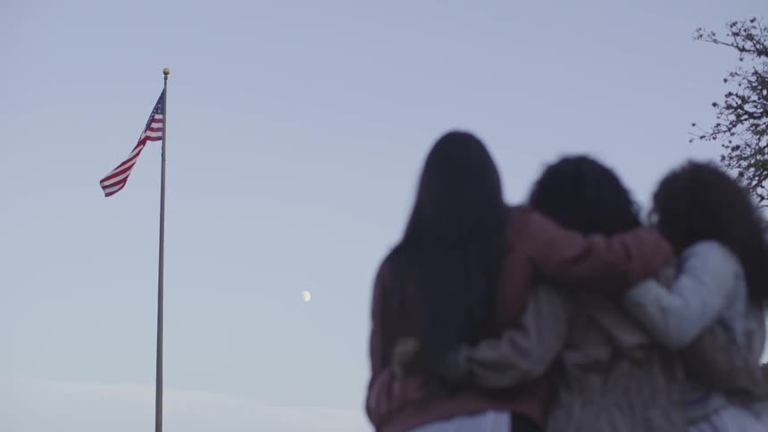 The young women look up at a flag blowing in the breeze at the top of a flag pole