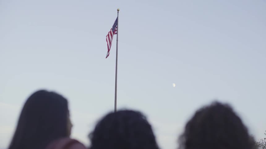 Three people standing below an american flag on a pole with the moon off to the side during the day