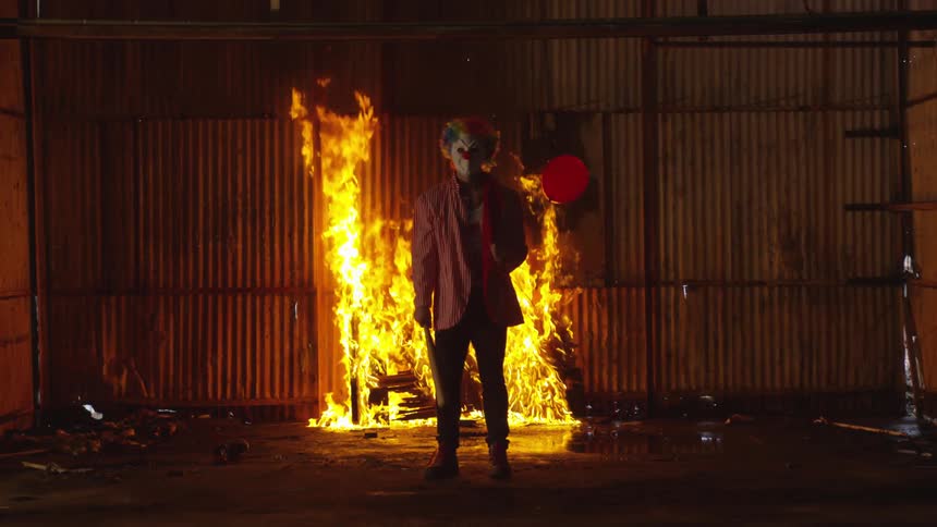 A clown stands in front of a fire while holding a baseball bat and red balloon