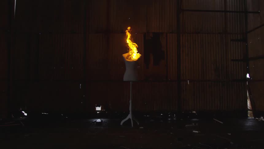 A top portion of a mannequin on a stand is on fire in a room