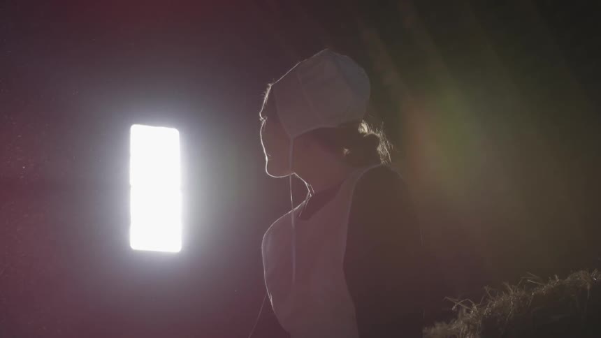 Young amish woman in bonnet sits in barn and looks towards window