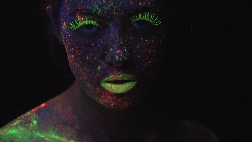 A woman covered in paint splatter poses under black light