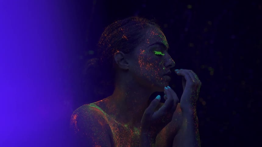 A woman covered in paint splatter poses under black light