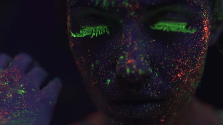 A person covered in neon makeup moves hand across their face