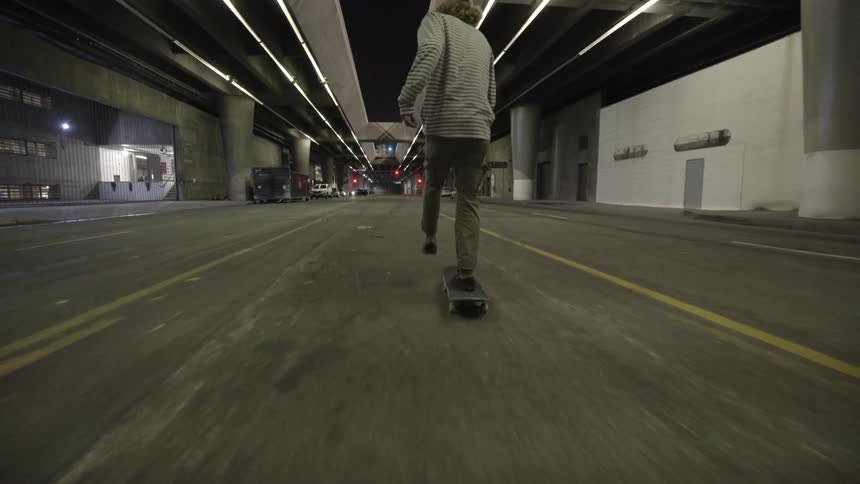 A young man rides a skateboard down the middle of a street beneath a structure