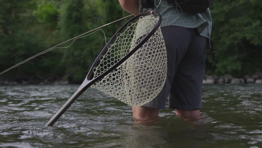 A man stands in a river fishing with a net clipped to his pack