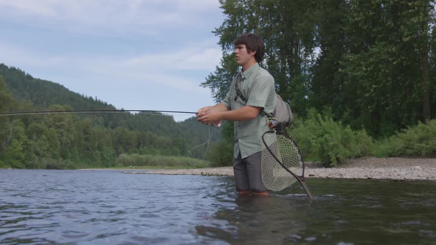 A man wearing shorts stands in a river and casts a fishing line
