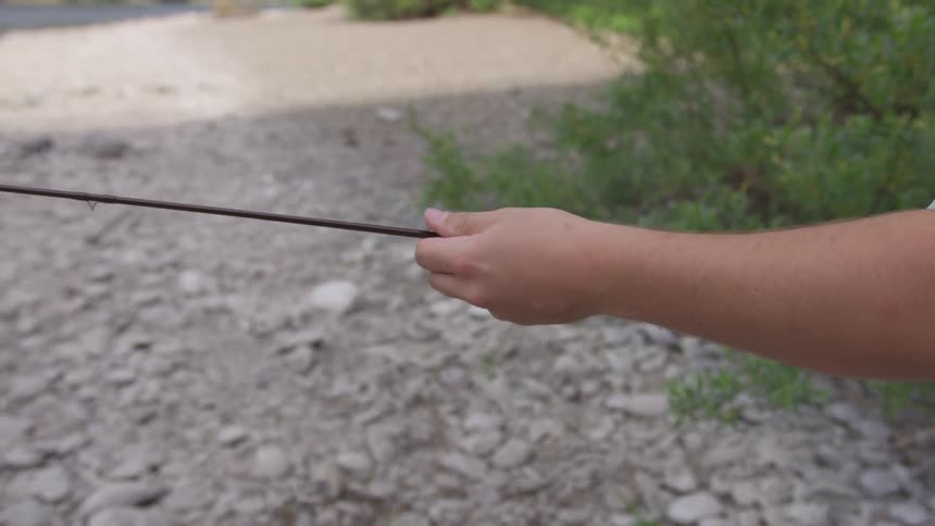 A man in a blue plaid shirt puts together a fishing rod near the edge of a river