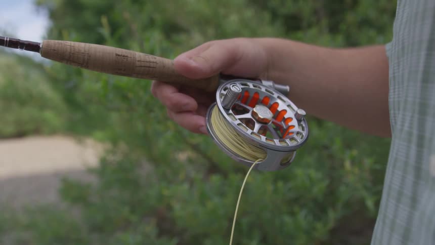 A man manually pulls the line from a fishing reel with trees in the background