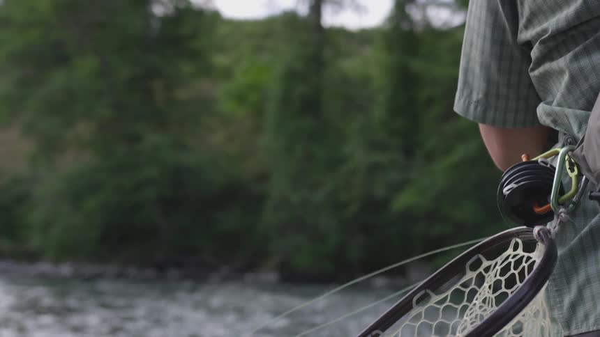 A man is at a briskly flowing river and he swings his fishing rod back and forth before casting the fishing line out a very far distance away from him into the river