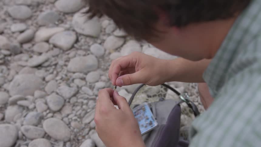 A young man kneeling in the rocks is tying lure to a fishing line