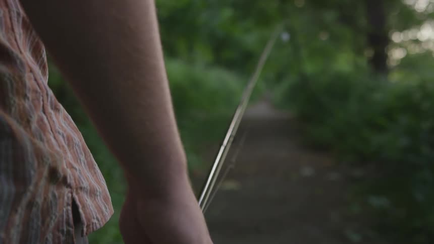 A man wearing a checkered shirt is walking on path in the woods while carrying a fishing rod