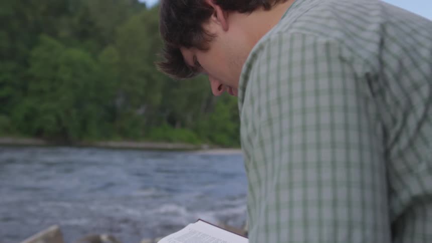 White man reading a book sitting on the rocks next to a river