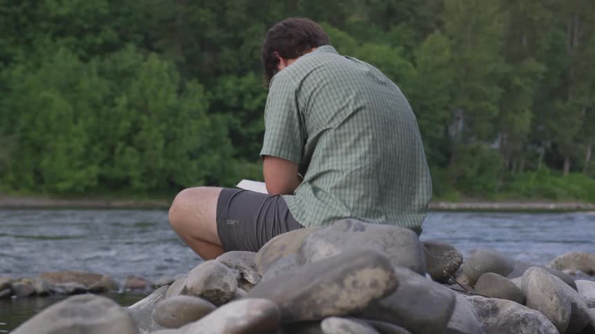 A man in shorts and a short sleeved shirt sits while reading a book on the rocky bank of river that is passing through a forested area
