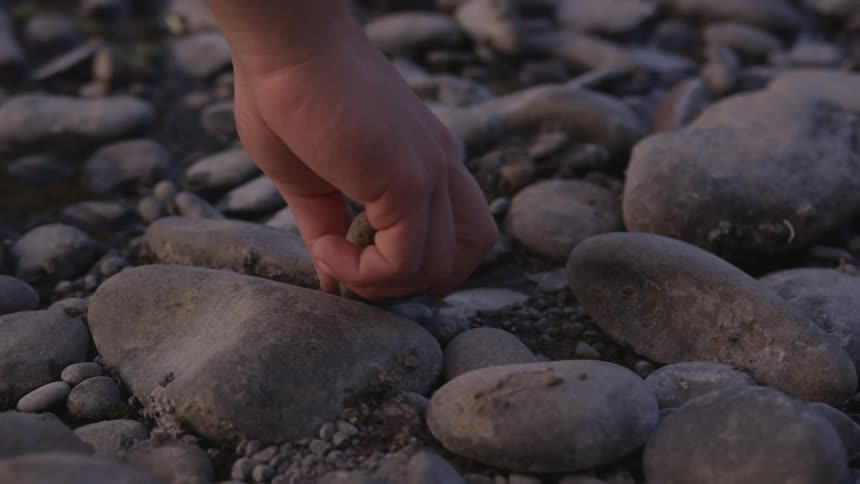 A hand reaches down and picks up a stone from a river bank