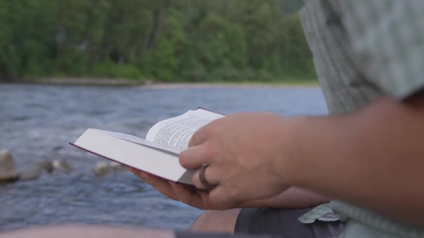 White man reading a book next to the river