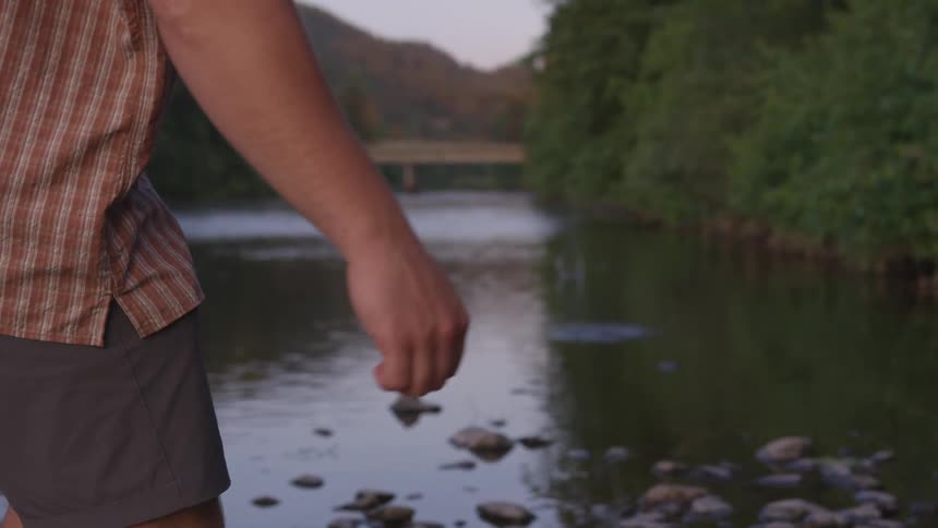 A male wearing a checkered shirt and khaki shorts is standing along the bank of the river skipping rocks into the water