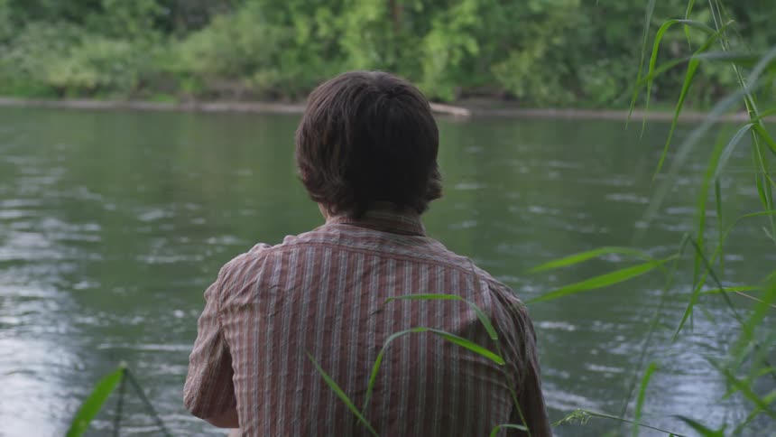 A male is sitting in the grass on the river bank watching the water flow