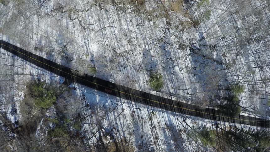 A bird's-eye-view of a car driving on a curved road through a wooded, rural area covered with snow