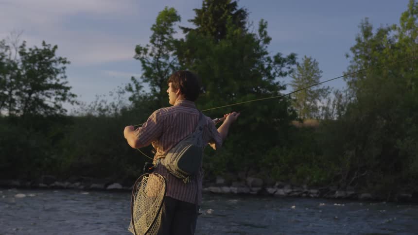 A young man stands in a river and casts his fishing line on a sunny day