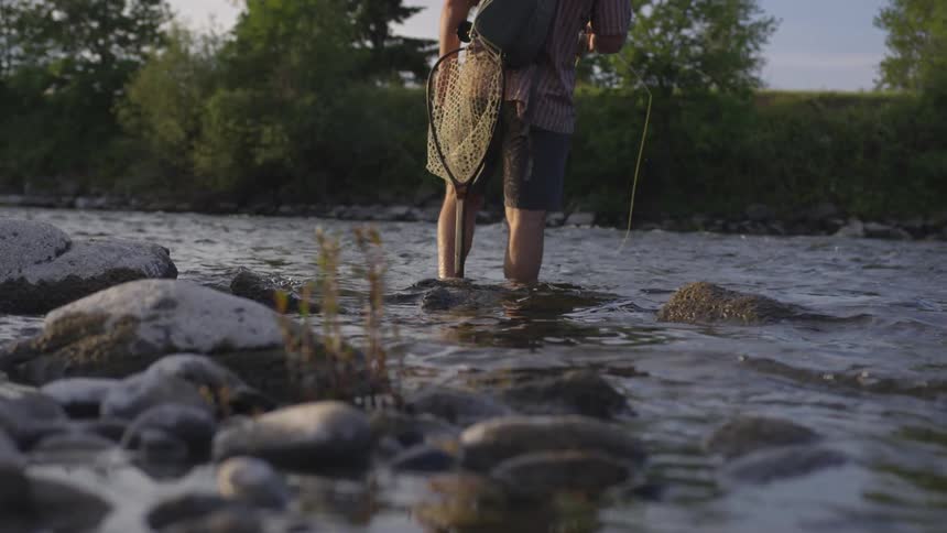 A man in shorts is standing in the stream beyond the rocks with a fishing net on his side and his line in the water