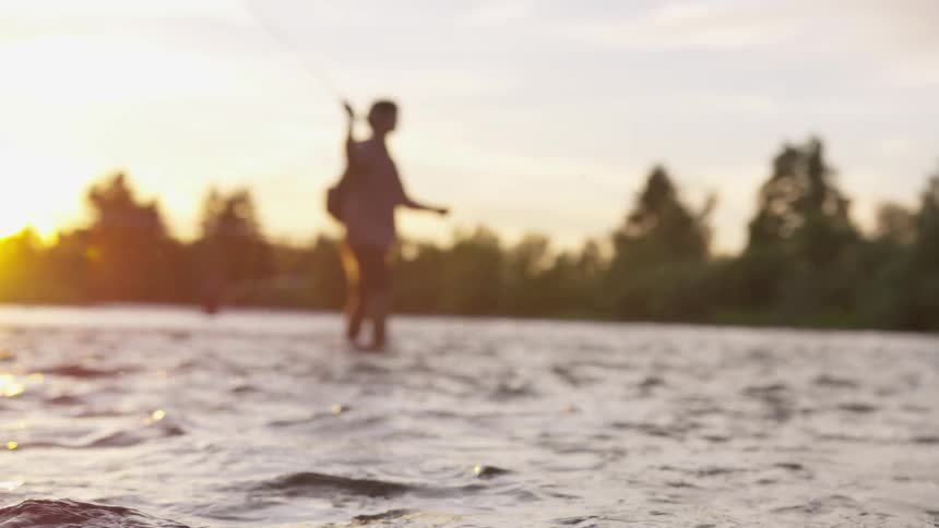 A man standing in ankle deep water casts a fishing line in a river with the sun rising behind him
