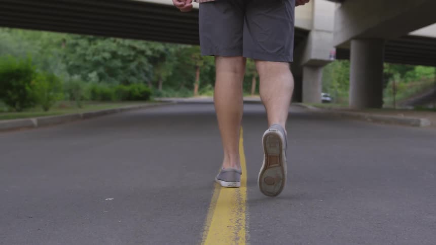 A man wearing gray shorts and light gray tennis shoes is walking along a yellow painted line in the middle of a road that passes underneath a large concrete overpass in an area with lots of trees