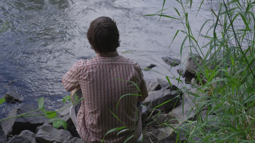 A male in a checkered shirt is kneeling on the rocks along the side of the flowing river