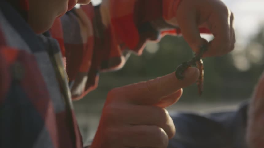 A young boy in a plaid shirt carefully picks up a dirt covered earthworm from the finger of an older man