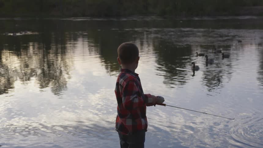 A boy reels in a fishing pole as ducks swim in the river in front of him