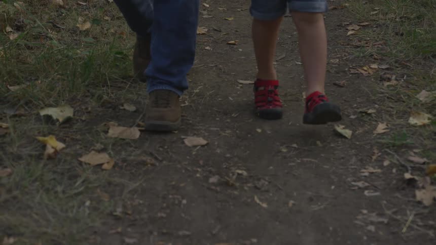 The feet of a young boy and a grown man are visible as they walk along a dirt path through a grassy area that is littered with fallen leaves