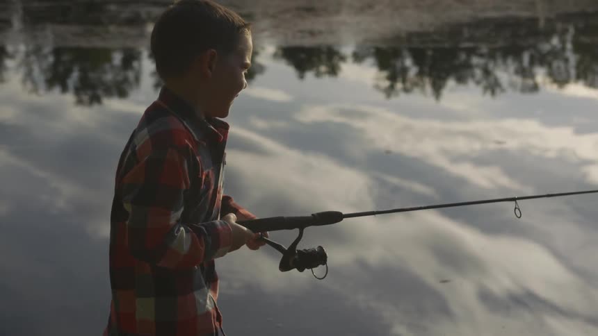 Boy turns reel of fishing rod while fishing by a lake