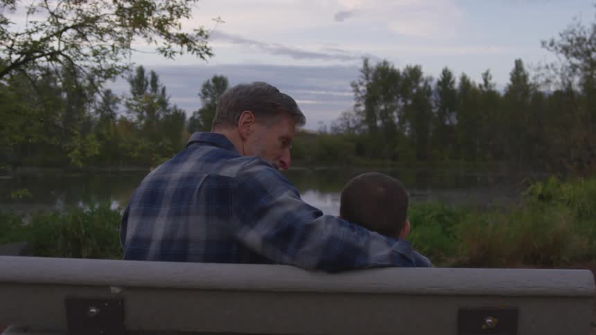 A dad sits on a lakeside bench with his arm around his son