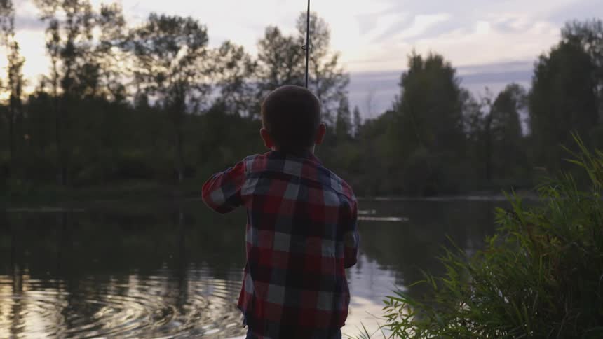 Young boy in a plaid shirt fishes at a lake at sunset