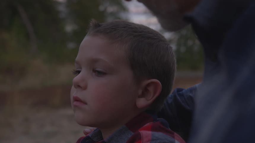 As they stand outdoors in low light an older man speaks to a young boy who is staring off into the distance