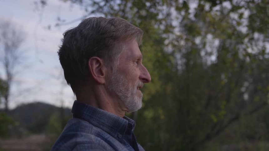 A older man with gray hair and a beard stands in a forested area and has a pleased look on his face