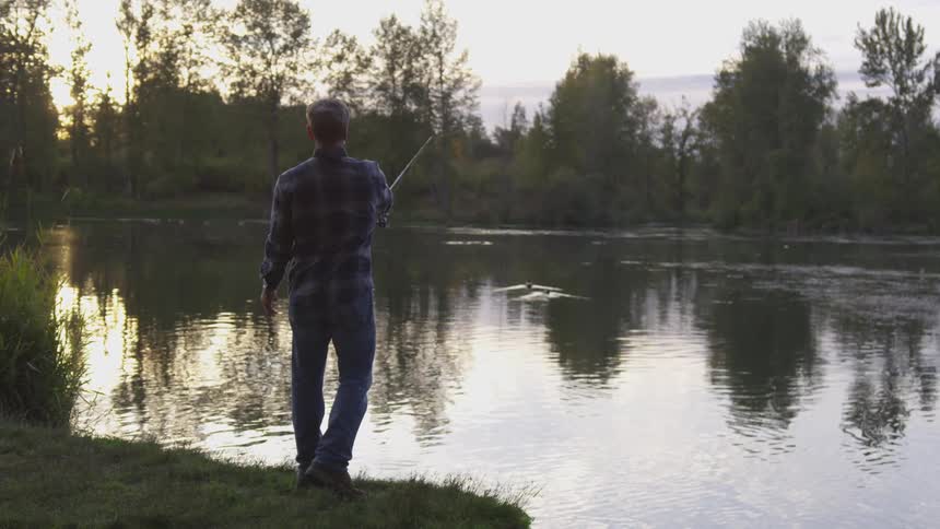 An older man in a blue flannel shirt stands at the edge of a lake and casts a fishing lure into the water by swinging the fishing pole