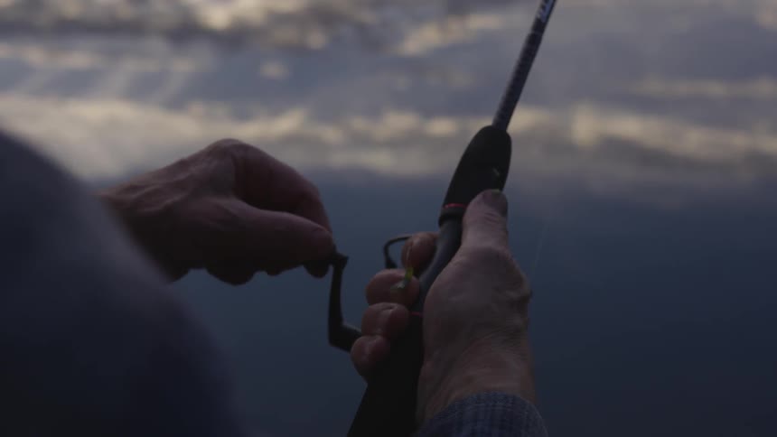 A man's hands turn the reel of his fishing rod and sky and clouds are reflected on the surface