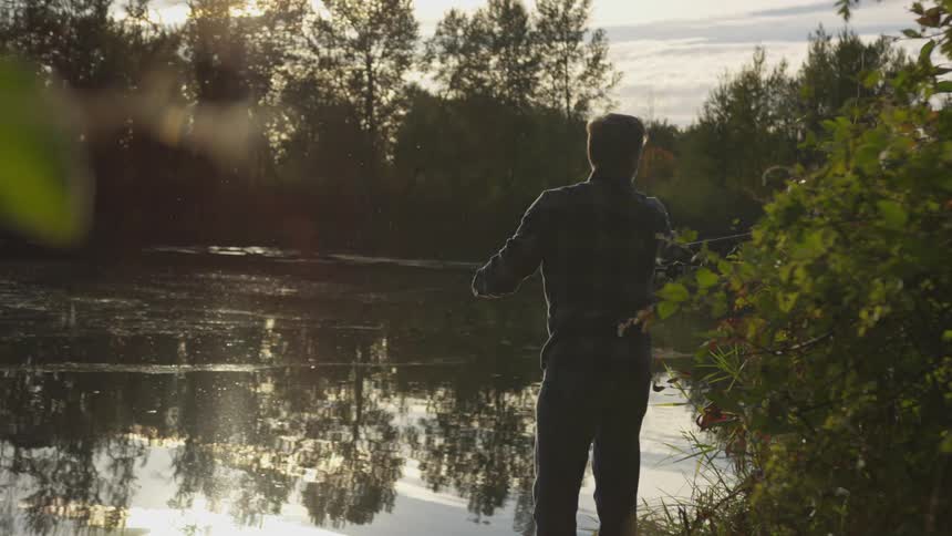 A man in a flannel shirt stands at the bank of a river with his fishing rod and prepares to cast his fishing lure